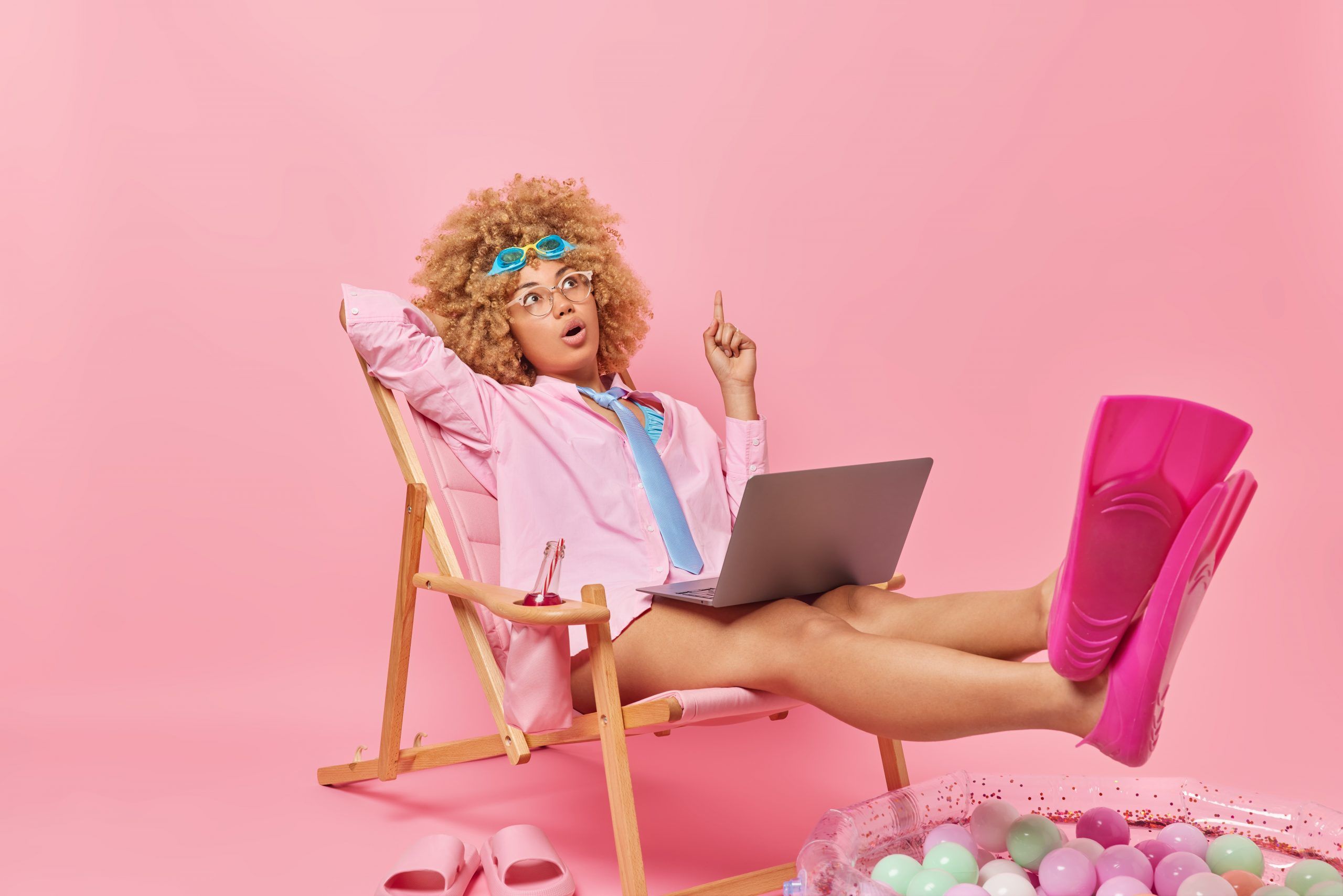 Surprised curly haired woman concentrated above points finger overhead dreams about summer vacations blogs in social networks using laptop poses on chaise lounge wears shirt tie and flippers